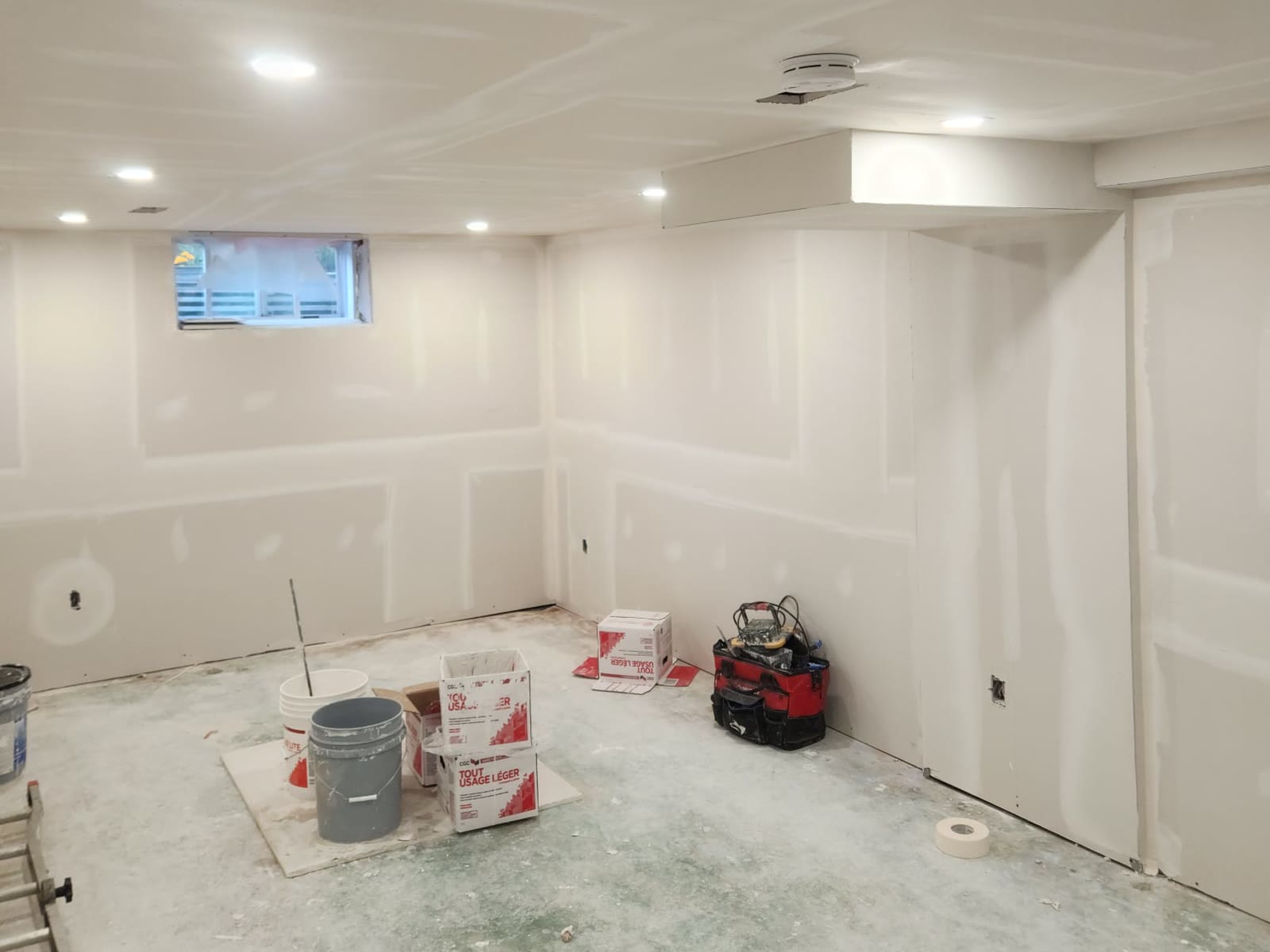 Basement room with taped and mudded drywall on the walls, ceiling and bulkhead, mud boxes and buckets on the floor