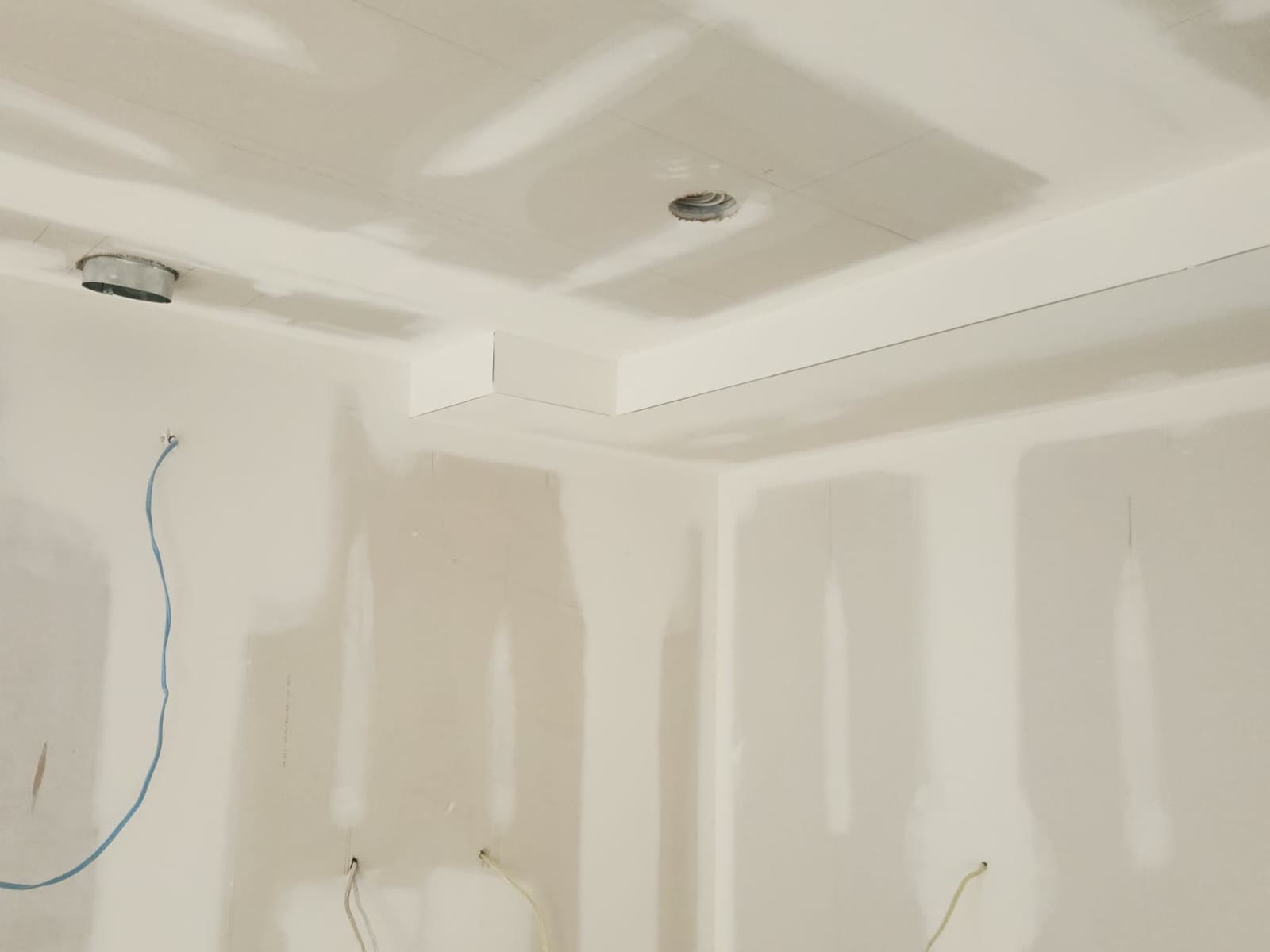 Room corner with a drywall bulkhead and taped, mudded ceiling and wall joints, electrical wires hanging for fixtures